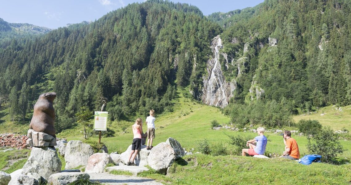 Family observing a marmot statue with a waterfall in the background. | © Holiday Region National Park Hohe Tauern